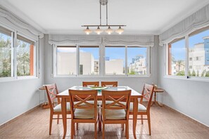 A dining room with a table and chairs, large windows, and a ceiling light fixture.
