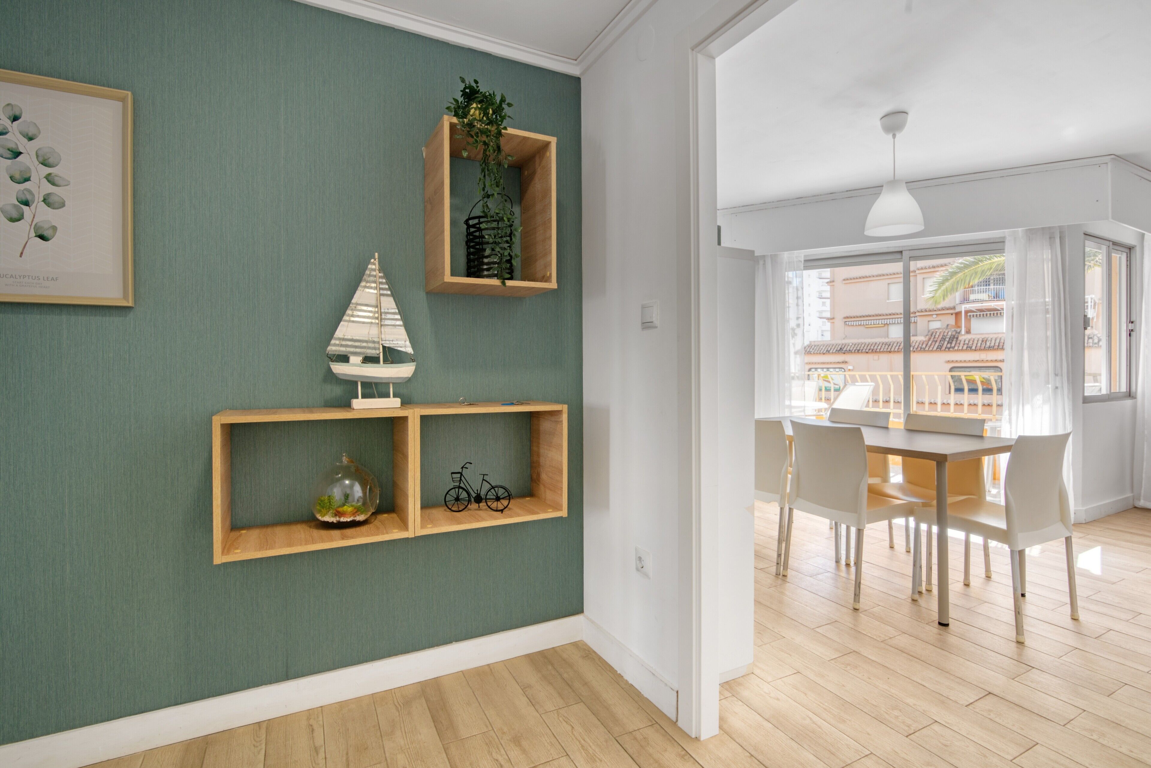 Una habitación con pared verde, estanterías de madera con adornos, una zona de comedor con sillas blancas y vistas a edificios a través de amplios ventanales.