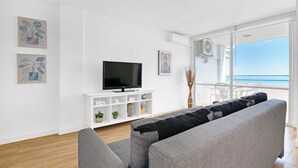 A modern living room with a grey sofa, a flat-screen TV on a white console, and a large window with a view of the sea. - Apartment 'Playa Puig' with Shared Pool, Wi-Fi and Air Conditioning (El Puig de Santa Maria)