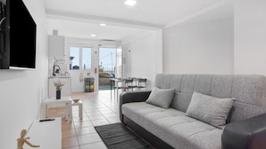 A modern living room with a grey sofa, a flat-screen TV mounted on the wall, a small table with a vase of flowers, and a kitchen area in the background. - Studio Apartment 'Babosas Charm' with Mountain View, Wi-Fi and Air Conditioning (Funchal)