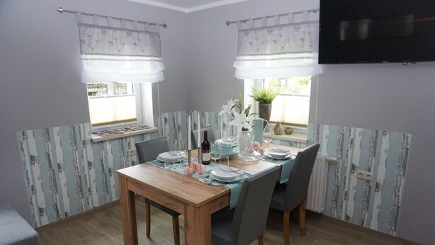 Un coin repas avec une table en bois dressée pour quatre personnes, entourée de chaises grises. On y trouve une fenêtre avec un rideau, une plante en pot et un téléviseur à écran plat fixé au mur.