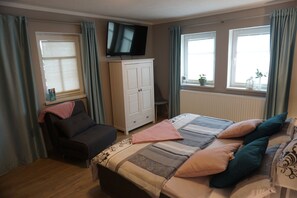 Quarto com cama, televisão, guarda-roupa e duas janelas com cortinas.