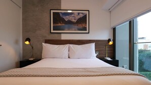 A modern hotel room with a large bed, two bedside lamps, a framed picture of a mountain landscape, and a large window offering a view of the outdoors.
