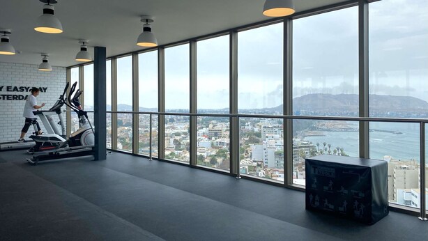 A gym with large windows, exercise equipment, and a view of a city and coastline.