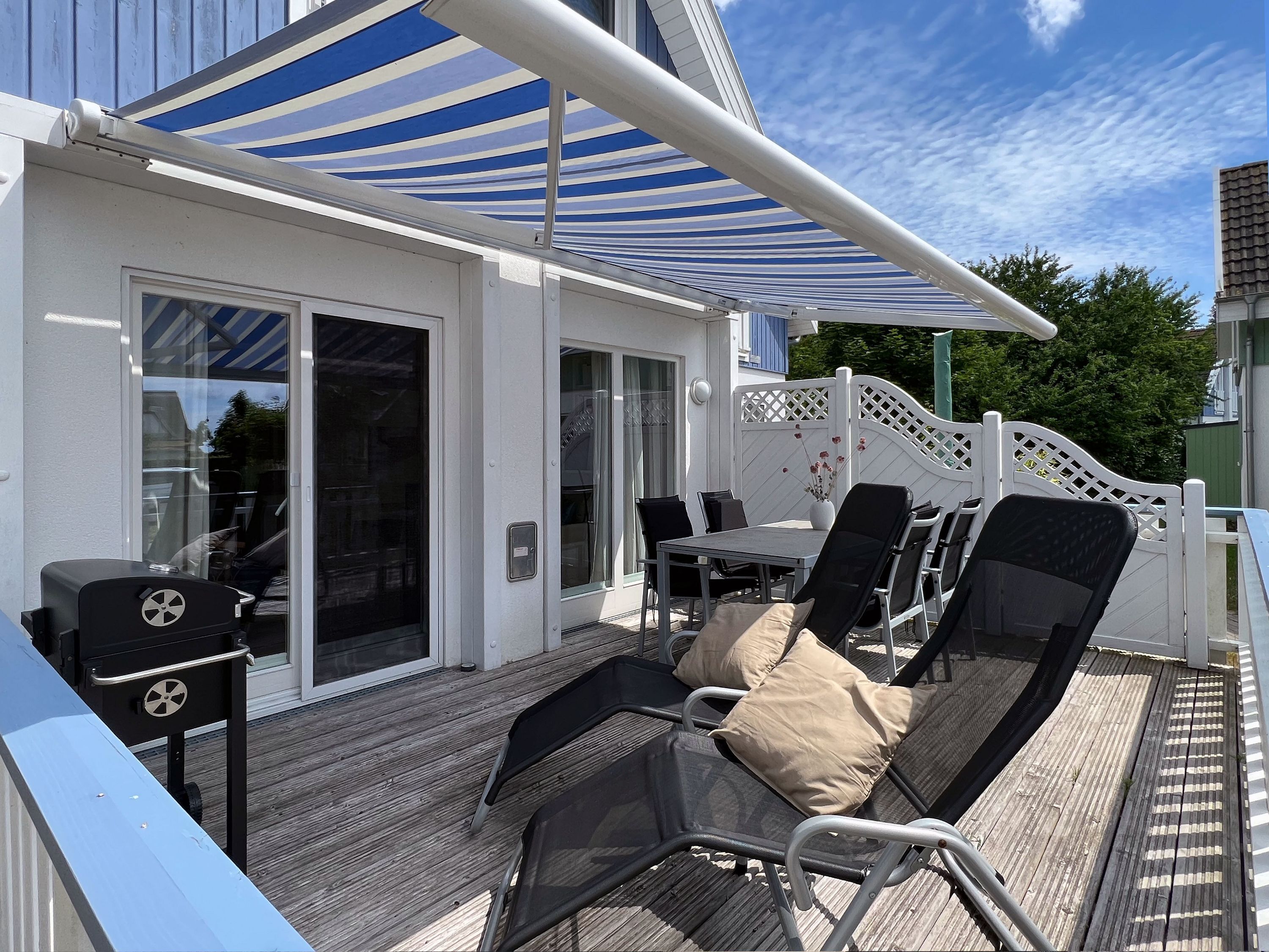 Een overdekt terras buiten met een tafel, stoelen, een barbecue en een witte schutting.