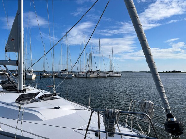 Un velero amarrado en un puerto deportivo con varios otros barcos, agua tranquila y cielo despejado.