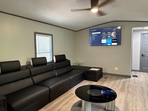 Un salon moderne avec un canapé d’angle, une table basse, un téléviseur à écran plat fixé au mur et une porte visible au fond.