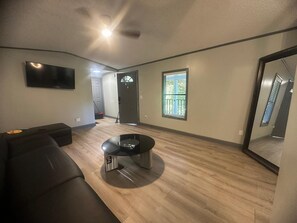 A modern living room with a flat-screen TV, a black leather sofa, a glass coffee table, and a sliding glass door leading to a balcony. - 2 bedroom renovated mobile home in Gainesville (Gainesville)
