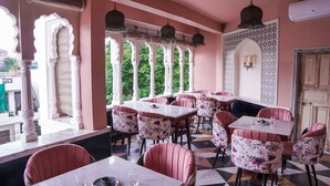 A restaurant with a pink and white color scheme, featuring traditional arches, patterned tiles, and a mix of floral and solid-colored chairs. Tables are set with plates and glasses. - Dev Mahal - A Boutique Heritage Hotel (Jaipur)