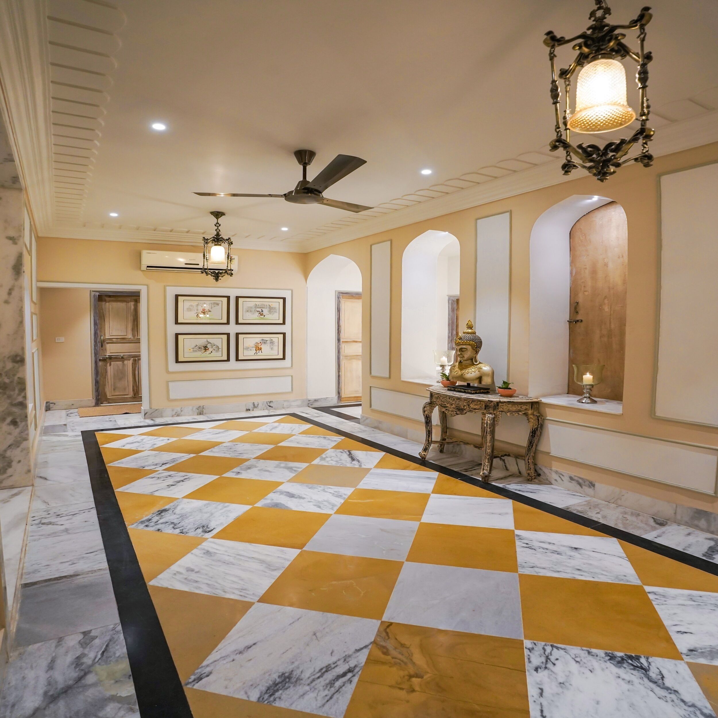 A spacious lobby with a checkered marble floor, a ceiling fan, wall-mounted air conditioning, framed artwork, and a decorative table with a statue.