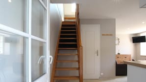 A modern interior with a wooden staircase leading to a second floor, a kitchen area with a wooden cabinet, and a white door. - Charming stone house with courtyard and terrace in Créances (Créances)