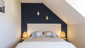 A bedroom with a sloped ceiling, a white headboard, two bedside lamps, and a bed with white bedding. - Charming stone house with courtyard and terrace in Créances (Créances)