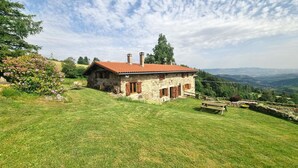 A stone house with a red roof, surrounded by a grassy hill and trees, overlooking a valley.