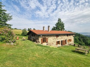 A stone house with a red-tiled roof, wooden shutters, and chimneys, surrounded by a grassy hill and trees. - Cozy cottage in the heart of Pilat with stunning views (Doizieux)