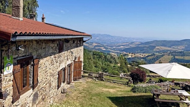 A stone building with a red roof, wooden shutters, and a view of rolling hills.