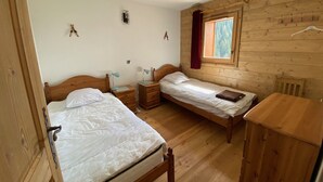 Une chambre avec deux lits en bois, une commode en bois et une fenêtre donnant sur des arbres.