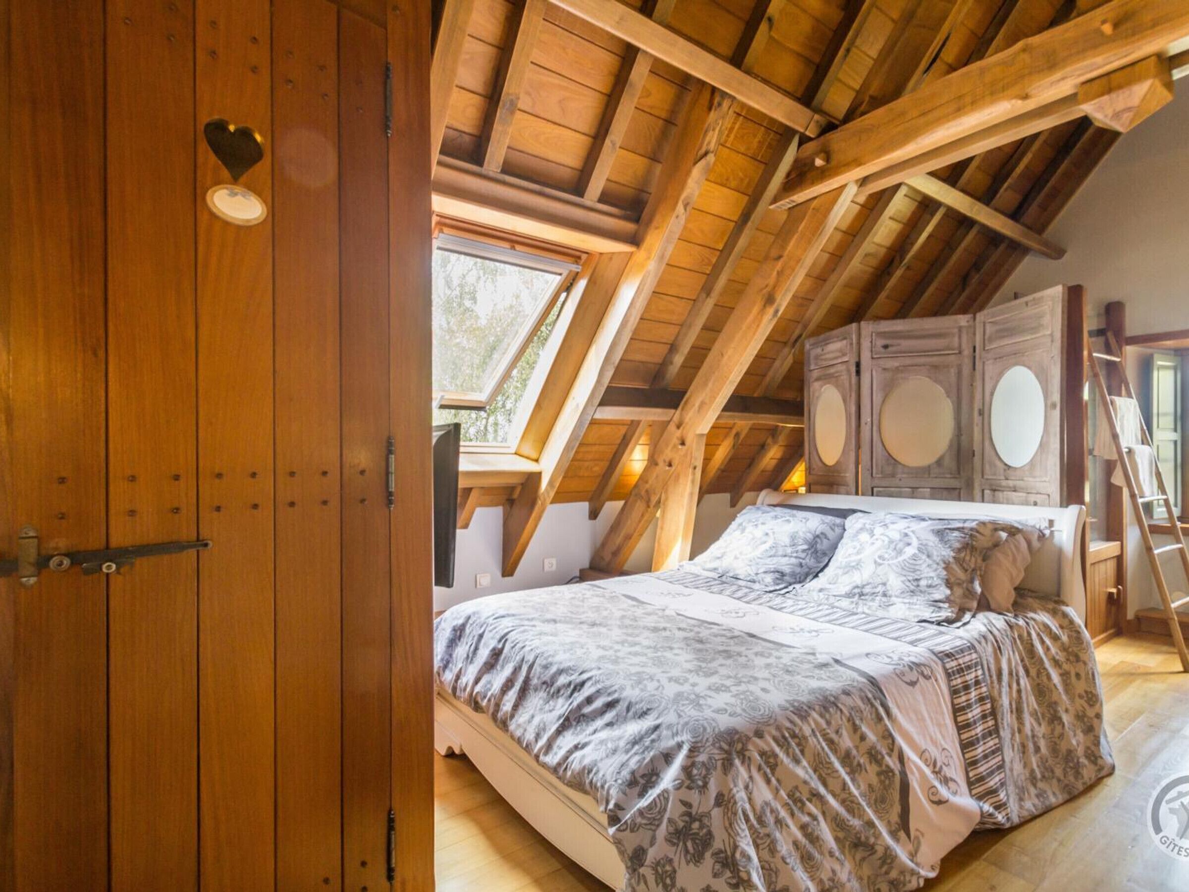 A bedroom with a sloped ceiling, wooden beams, a bed with floral bedding, a wardrobe with round mirrors, and a skylight.