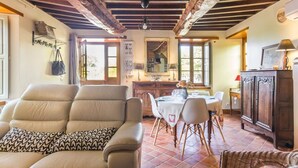 A cozy living room with a sofa, dining table, chairs, and a wicker chair. There is a kitchen area with a stove and cabinets. - Charming stone house with garden, ideal for couple (Gavray-sur-Sienne)