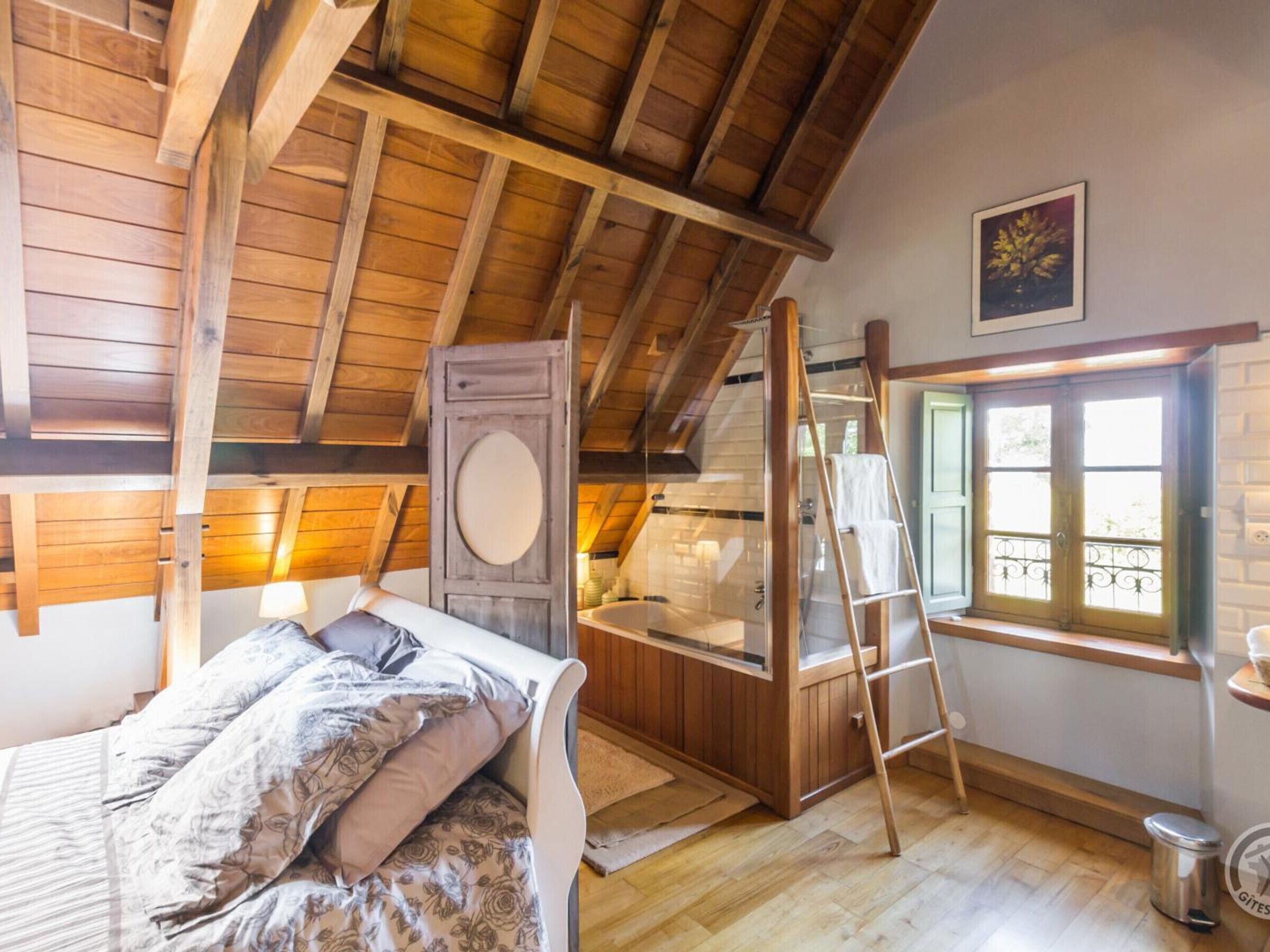 A cozy attic bedroom with a wooden ceiling, a bed with floral bedding, a large window with a view, and a ladder leading to a lofted space.