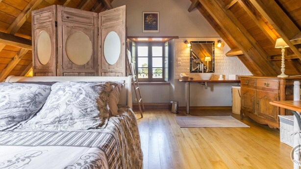 A cozy attic bedroom with a wooden ceiling, a large bed with floral bedding, a wooden wardrobe, a bathroom with a mirror and sink, and a window letting in natural light. - Charming stone house with garden, ideal for couple (Gavray-sur-Sienne)
