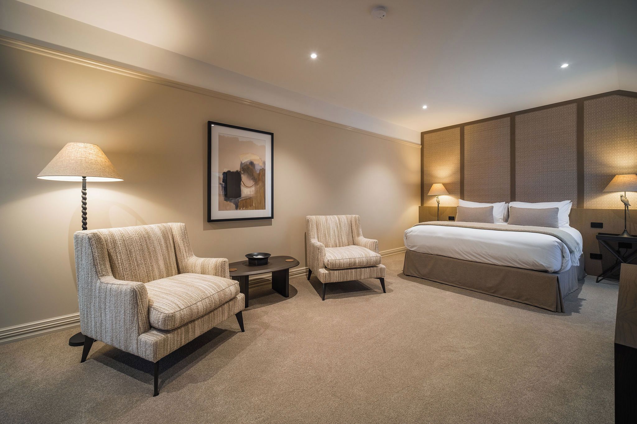 A hotel room with a large bed, two armchairs, a small table, and a framed picture on the wall.