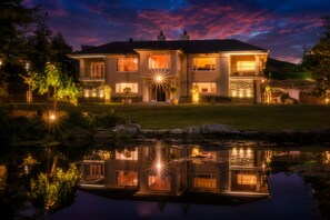 Een groot, twee verdiepingen tellend huis met een verlichte veranda en een vijver op de voorgrond.
