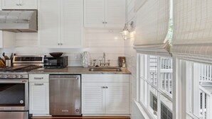 A modern kitchen with stainless steel appliances, white cabinetry, and a large window with white blinds. - 'Garden Cottage' at The Broad Street Residences (Charleston)