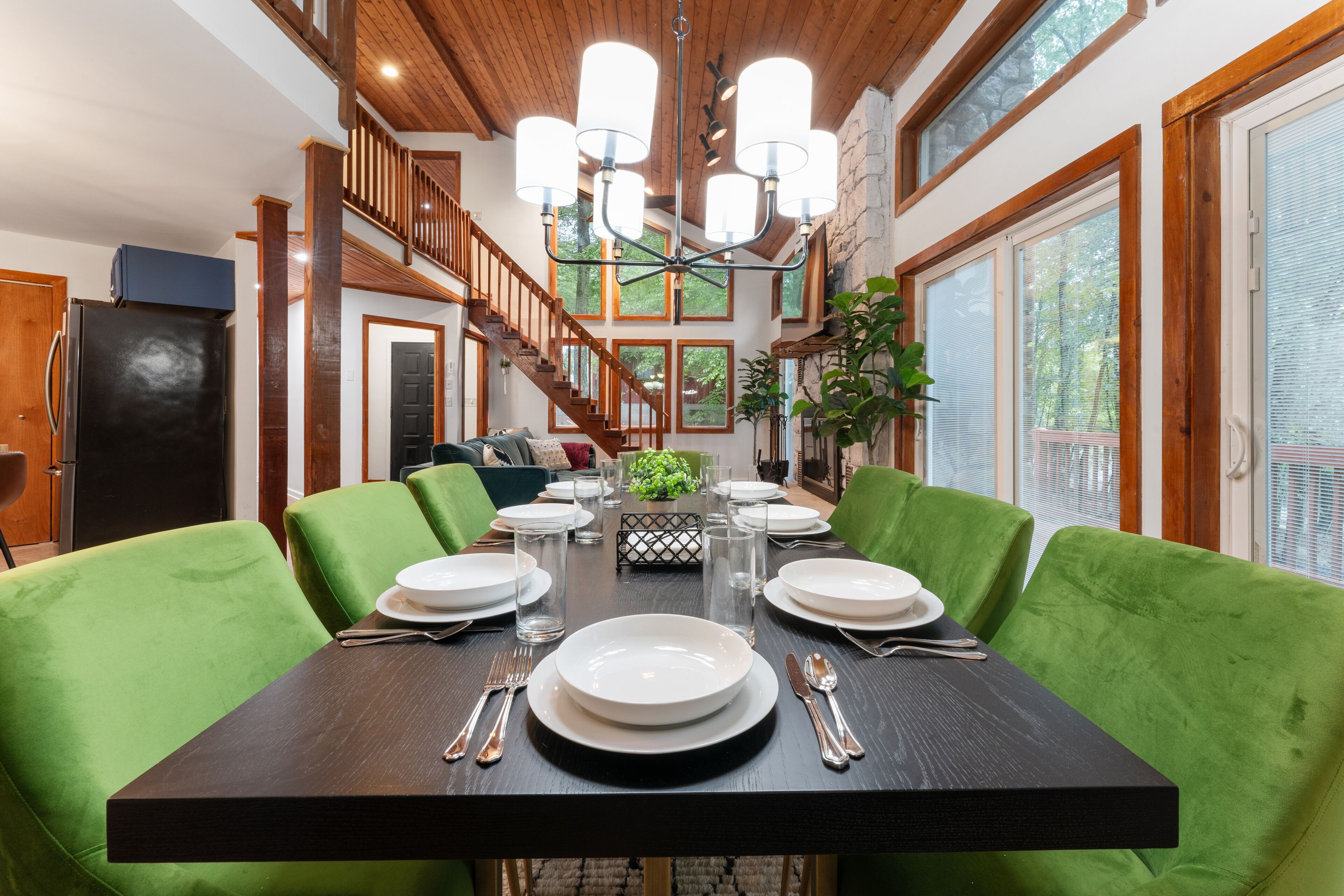 A dining area with a dark wooden table set for six, surrounded by green upholstered chairs. A chandelier hangs from the ceiling, and a staircase with wooden railings is visible in the background.