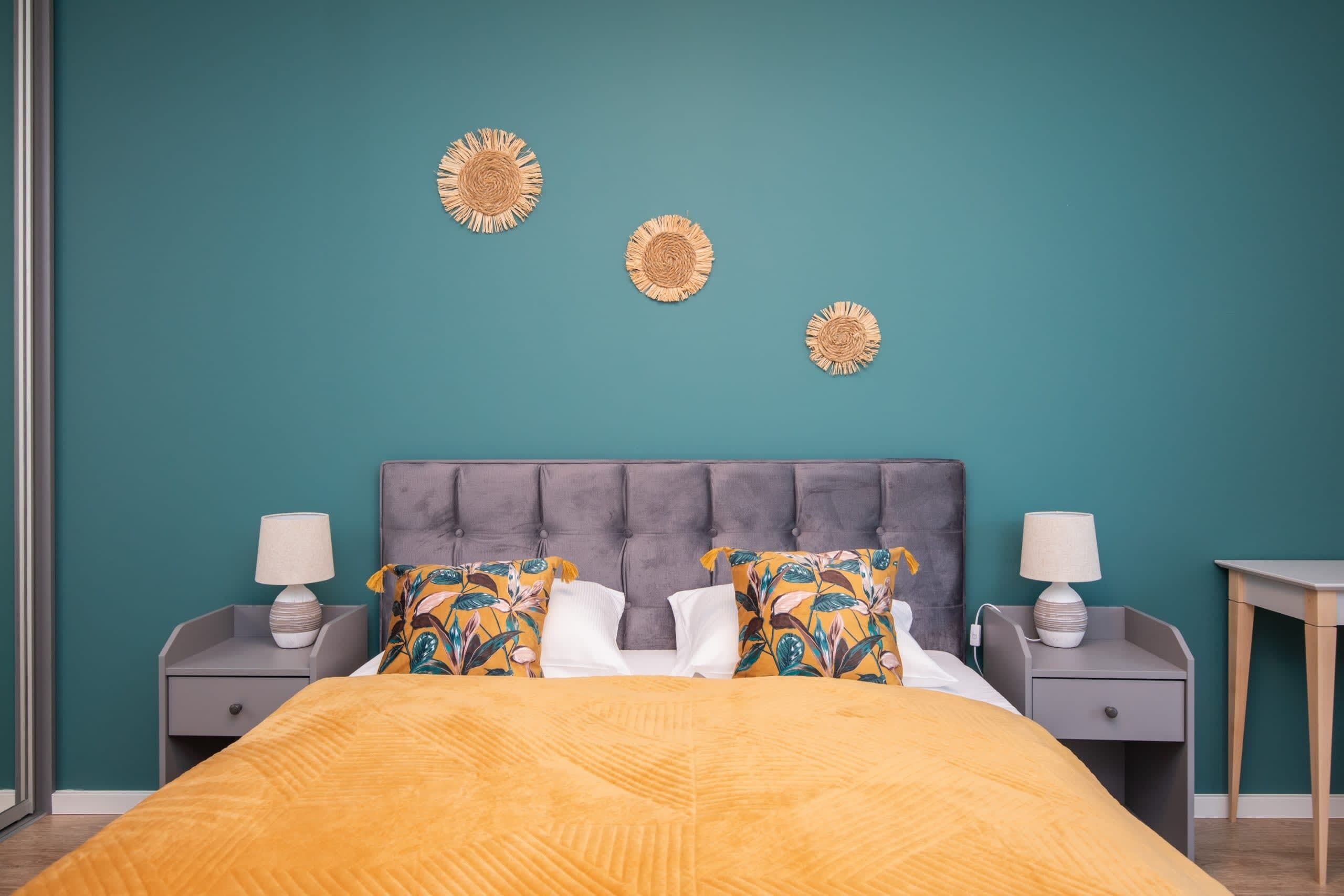 A bedroom with a teal wall, a tufted headboard, a bed with a yellow blanket, two nightstands with lamps, and decorative wall art.