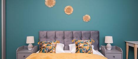 A bedroom with a teal wall, a tufted headboard, a bed with a yellow blanket, two nightstands with lamps, and decorative wall art.
