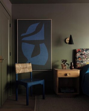 Una habitación con una silla azul, una mesita de noche de madera con una lámpara y un cuadro abstracto grande en la pared.