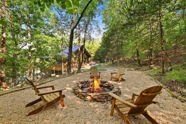 A forested campsite with a stone fire pit, wooden chairs, and a cabin in the background.