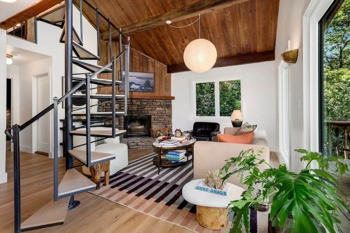 A modern living room with a stone fireplace, a wooden staircase, and a large window overlooking greenery.