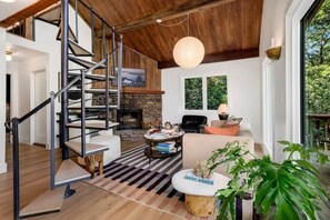 A modern living room with a stone fireplace, a wooden staircase, and a large window overlooking greenery.
