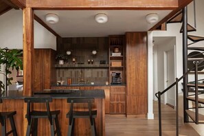 A modern kitchen with wooden cabinetry, a central island with bar stools, and a staircase with a metal railing. - Canyon Grove - Secluded lake retreat (Garfield)