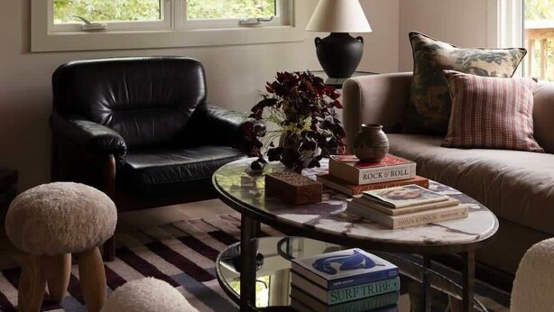 A living room with a black leather sofa, a round coffee table with books, a lamp, and a large window with a view of greenery.