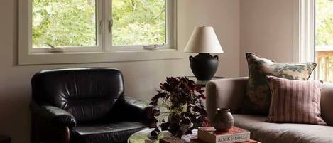 Un salon avec un canapé en cuir noir, une table basse ronde sur laquelle sont posés des livres, une lampe et une grande fenêtre donnant sur un paysage verdoyant.