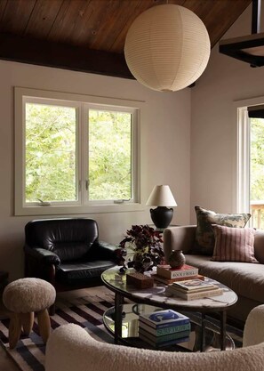 A cozy living room with a black leather sofa, a round coffee table with books, a lamp, and a large window with a view of greenery.