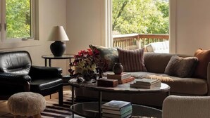 A cozy living room with a leather chair, a sofa, a coffee table with books, a lamp, and a view of greenery outside. - Canyon Grove - Secluded lake retreat (Garfield)