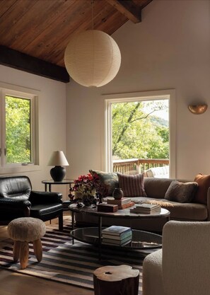 A cozy living room with a leather chair, a sofa, a coffee table with books, a lamp, and a view of greenery outside. - Canyon Grove - Secluded lake retreat (Garfield)