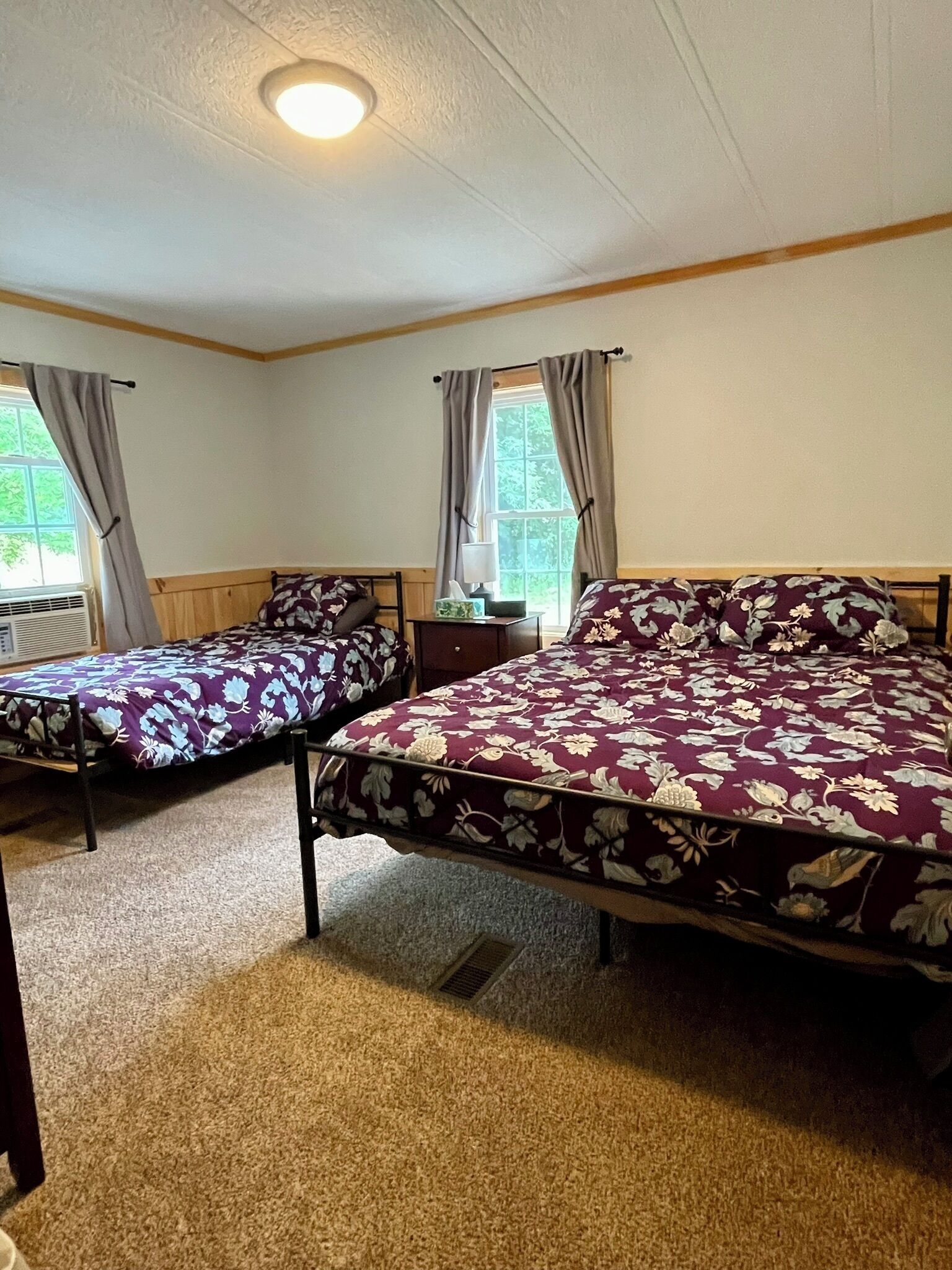 A room with two single beds and a larger bed, all with floral bedding. There is a window with curtains, a radiator, and a small table with a lamp.