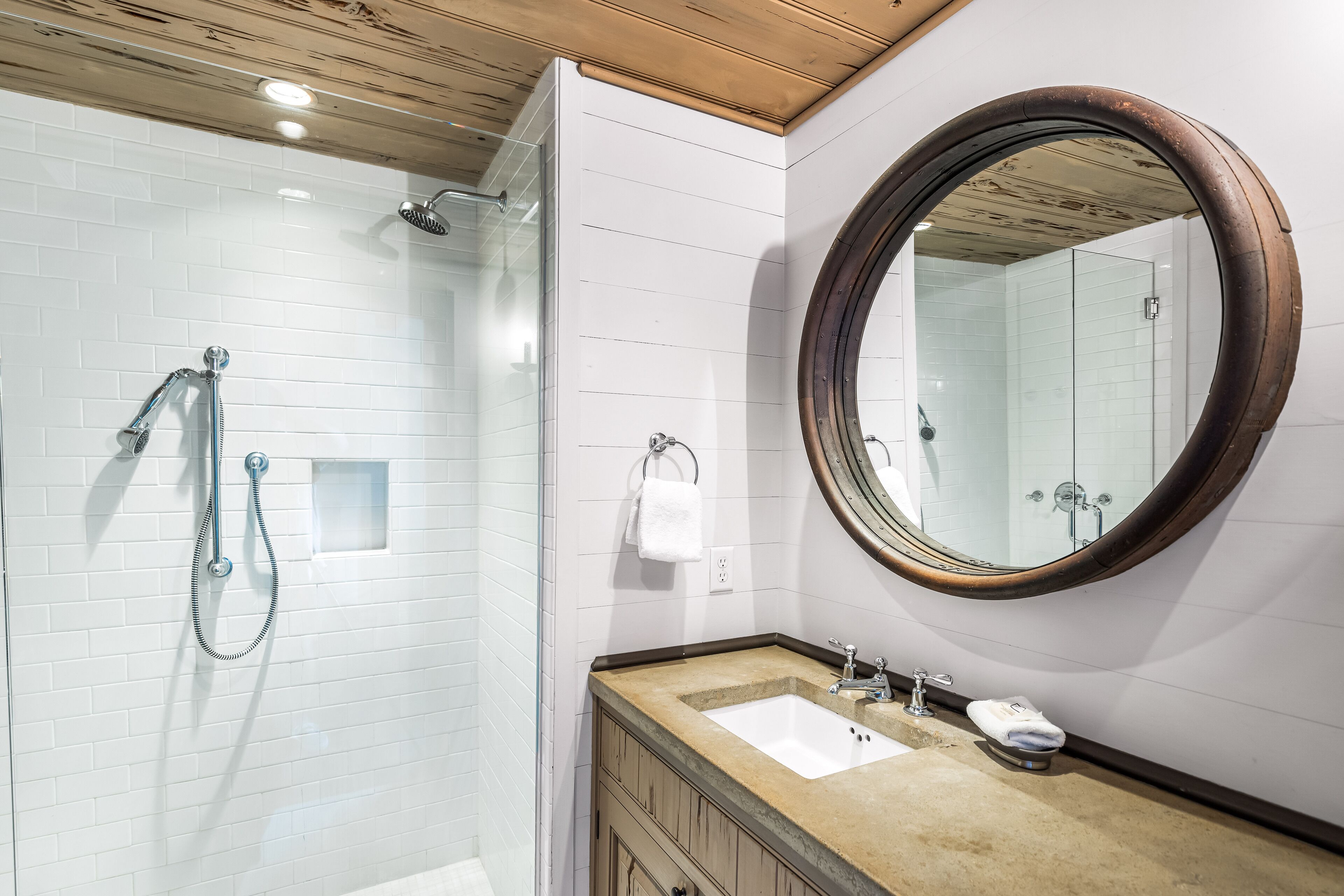 A bathroom with a glass shower enclosure, a wooden ceiling, a white tiled wall, a round mirror, a sink with a beige countertop, and a towel rack.