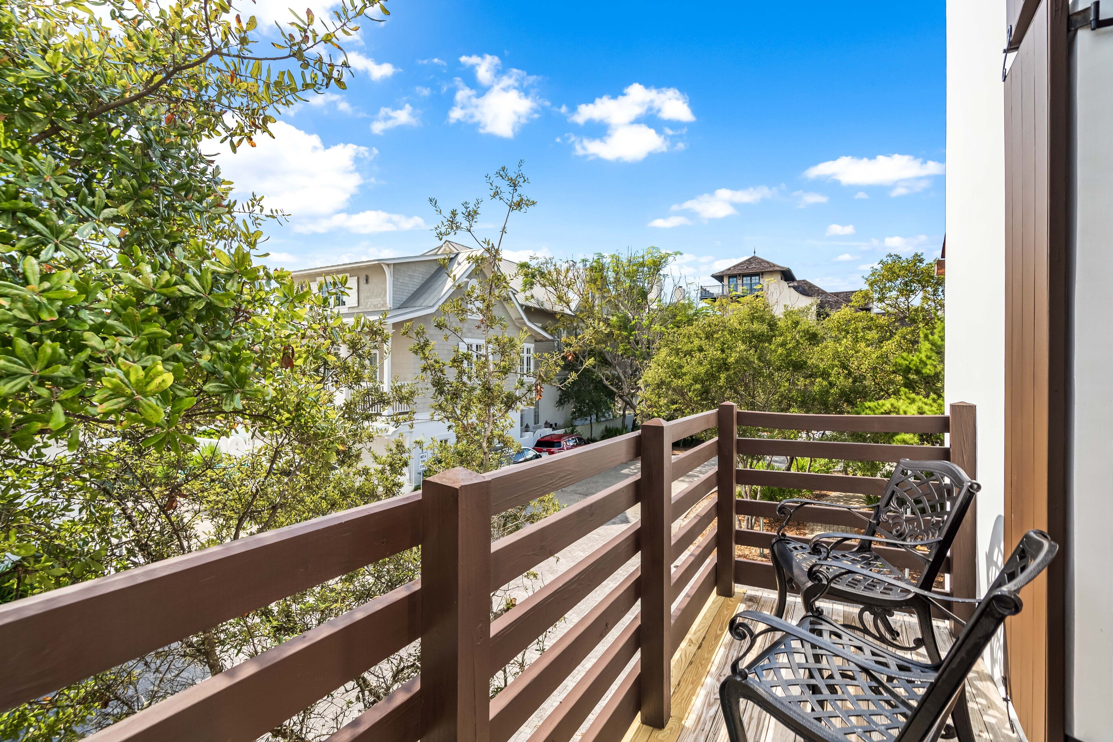 A balcony with wooden railings and two metal chairs, overlooking a residential area with houses and trees.