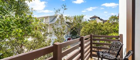 A balcony with wooden railings and two metal chairs, overlooking a residential area with houses and trees.
