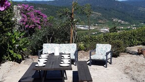 Ein Außenbereich zum Sitzen mit Tisch und Stühlen, umgeben von Grünpflanzen und Blick auf eine entfernte Landschaft.