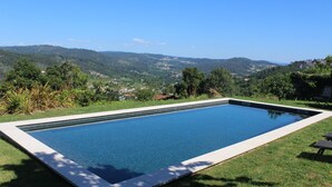 Ein rechteckiger Swimmingpool mit klarem, blauem Wasser, umgeben von einer Rasenfläche und Bäumen, mit Blick auf ein Tal und ferne Hügel.