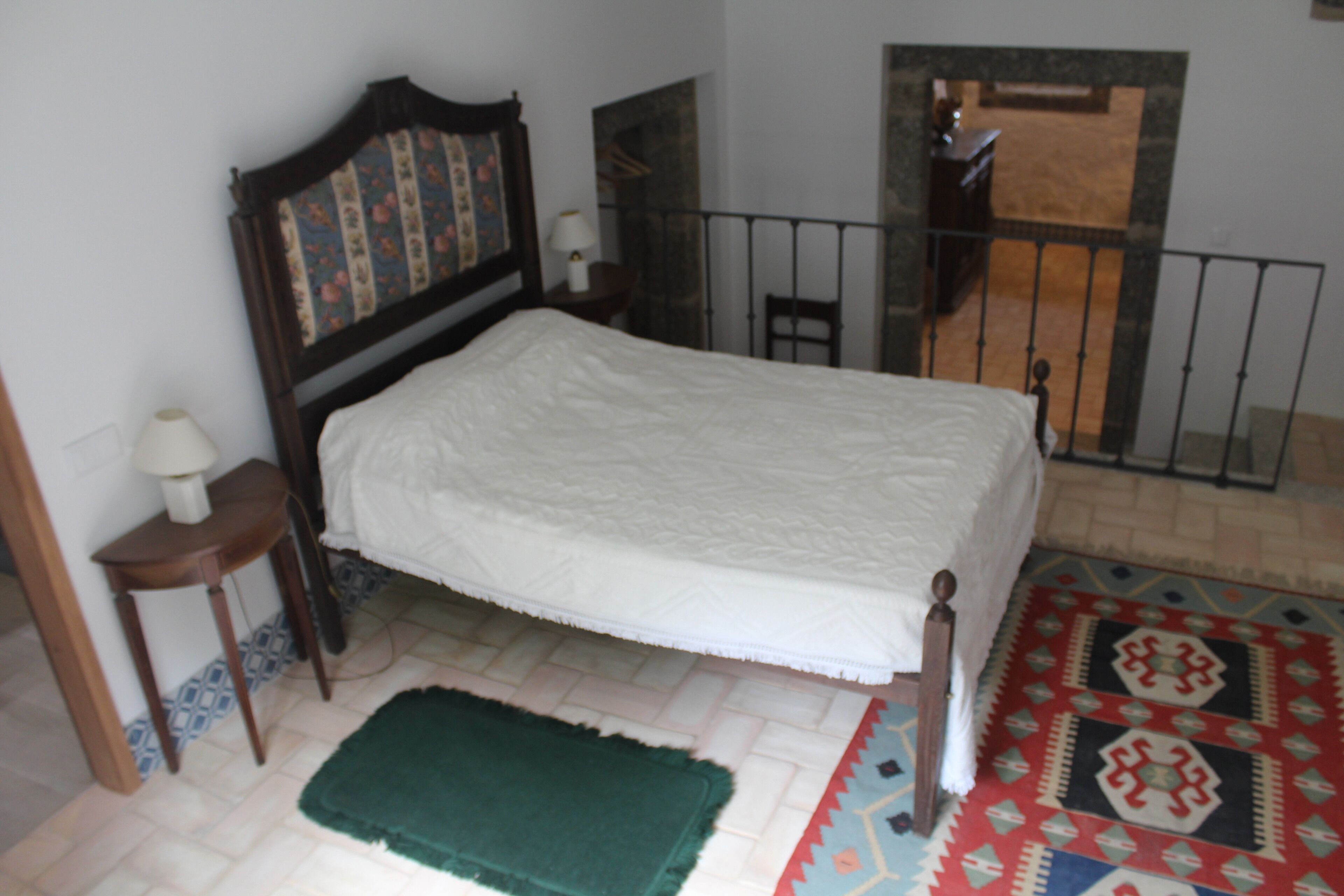 Ein Schlafzimmer mit einem Bett, einem Nachttisch, einer Lampe, einem gemusterten Teppich und Blick ins Freie durch ein Fenster.
