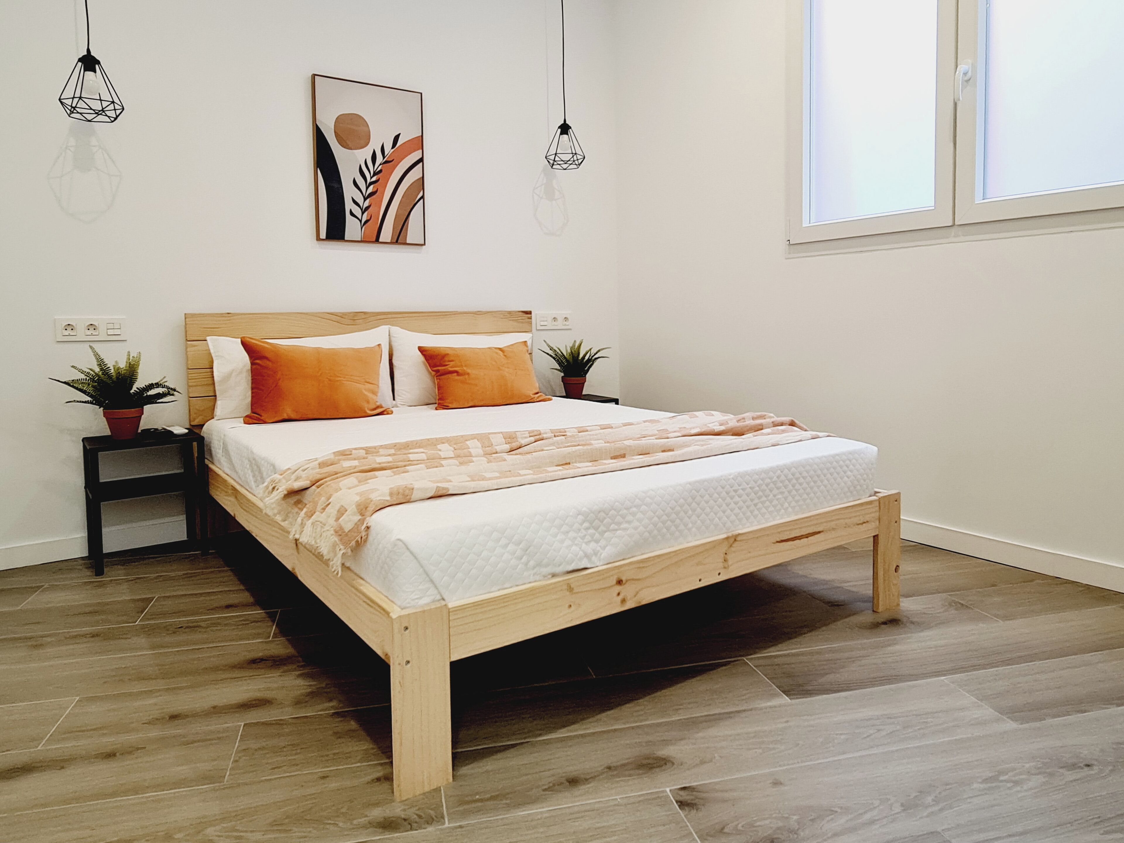 Un dormitorio con una cama de madera, dos mesitas de noche, una planta en maceta, una obra de arte colgada en la pared y una ventana con persianas.