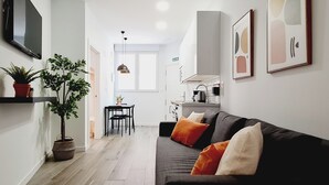 Een moderne woonkamer met een donkere bank, een eettafel en stoelen. Er staan potplanten op een plank en een flatscreen televisie is aan de muur bevestigd.