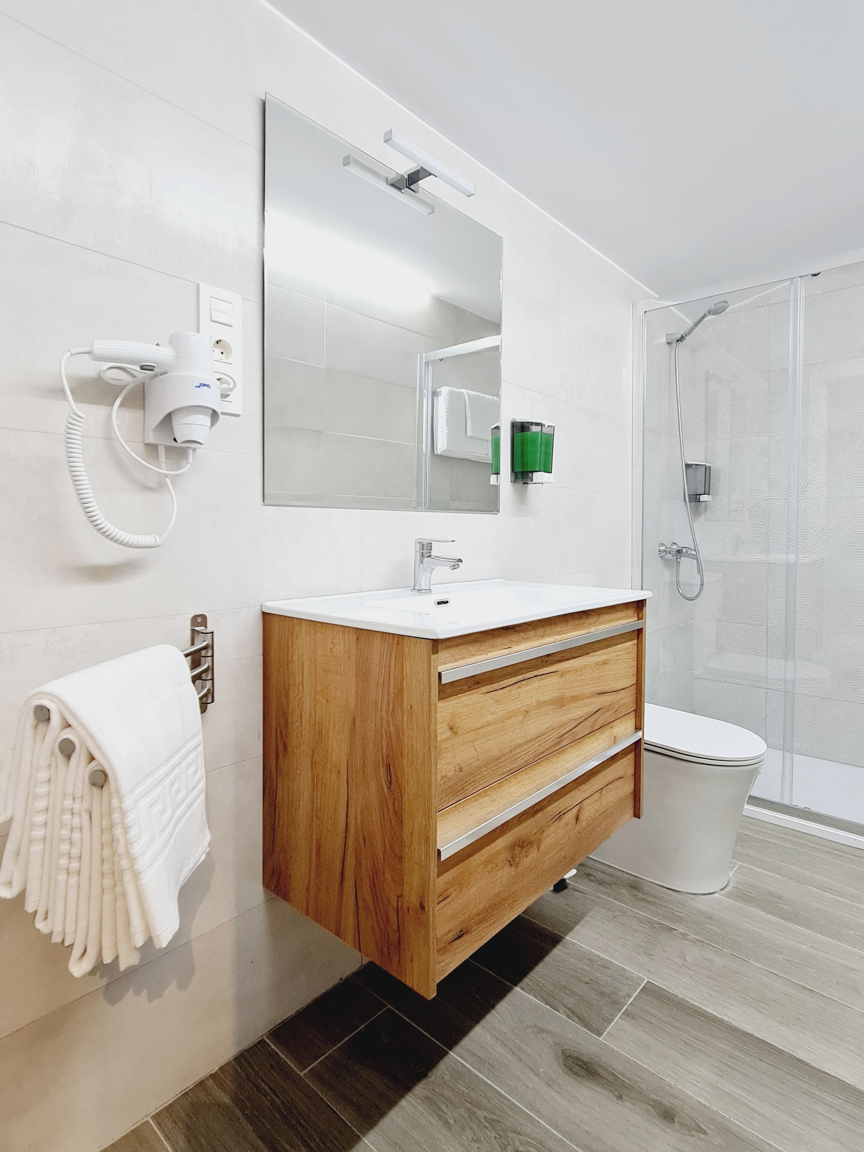 A modern bathroom with a wooden vanity, a large mirror, a white sink, a towel rack, and a glass shower enclosure.